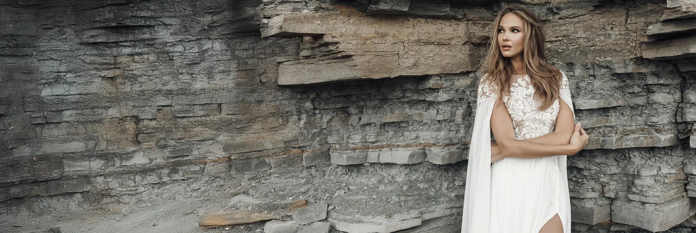 Eine Frau trägt ein weißes Spitzenhochzeitskleid mit Cap-Ärmeln und offenem Rücken, kombiniert mit einem fließenden Elfenbeincape.